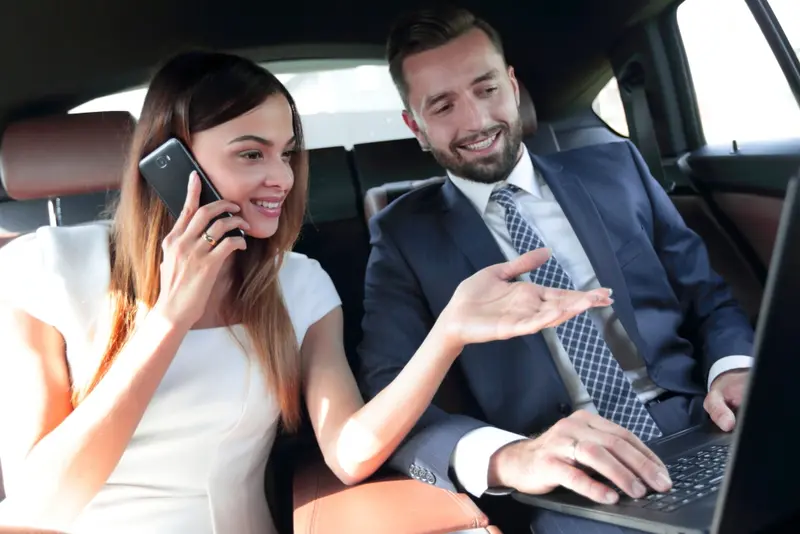 A businessman reviews documents on his laptop while a businesswoman beside him speaks on her phone, both seated in the back of a luxury black car, symbolizing professional Houston Hobby Airport to Galveston transfers.